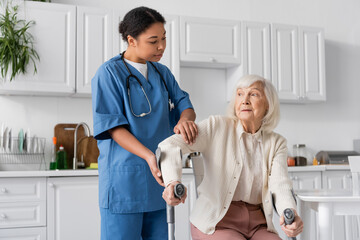 multiracial nurse in uniform helping senior woman using crutches to stand up in modern apartment.