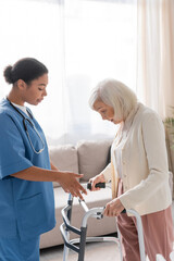 Fototapeta premium brunette multiracial nurse in blue uniform looking at senior woman with grey hair walking with help of walker at home.