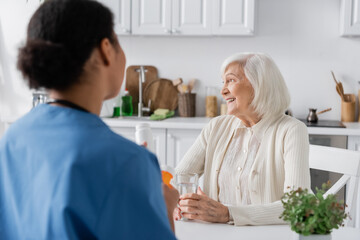 happy retired woman with grey hair looking at away near multiracial nurse on blurred foreground.