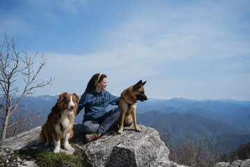 Happy young Caucasian woman sitting on large stone with two dogs and smiling. Snowy peaks of mountains are visible in distance. Traveling concept and hiking in mountains with dog.
