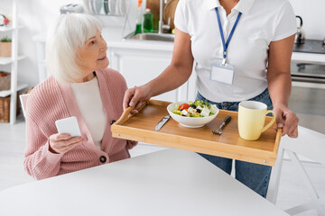 multiracial social worker holding tray with lunch for happy senior woman with grey hair.