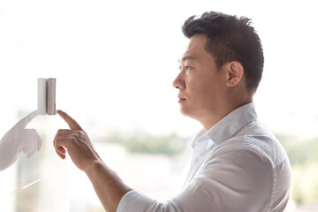 An Asian man stands near an electronic panel on the wall and presses the entry bell of an automatic door with his finger. Configures the wall conditioner, the security system. - Powered by Adobe