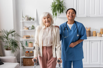 Fototapeta premium positive retired woman with grey hair using crutches while walking near happy multiracial nurse at home.