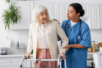 brunette multiracial nurse in blue uniform supporting senior woman with grey hair walking with help...
