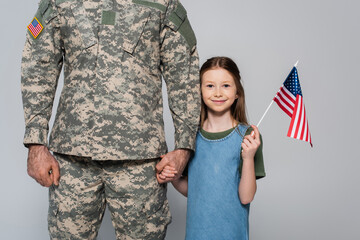cheerful preteen girl holding American flag and hand of father in military uniform during memorial day isolated on grey.