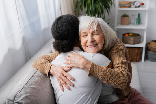 happy senior woman with closed eyes hugging curly social worker in living room.