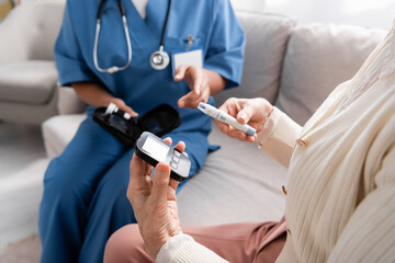 cropped view of senior woman holding lancet pen and glucometer near multiracial nurse sitting on...