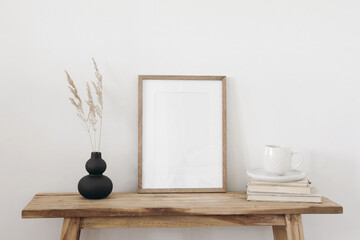 Blank vertical wooden picture frame mockup. Organic shaped black vase with dry grass on table,...
