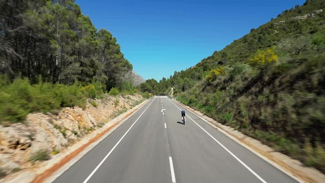 Aerial drone footage by a FPV drone shows a solo male cyclist training on a time trial bike on empty road preparing to competition.Triathlete riding very fast on tt bicycle.Alicante region in Spain
