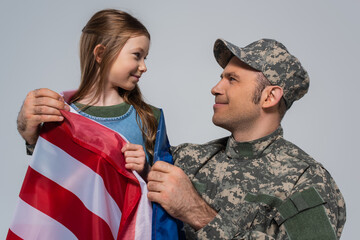 Fototapeta premium serviceman in military uniform looking at happy daughter wrapped in flag of United States of America isolated on grey.