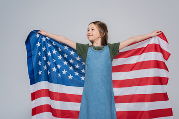 patriotic preteen girl smiling while holding huge flag of America during memorial day isolated on grey.