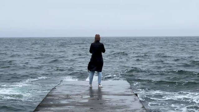 Back view of girl standing alone on pier near the sea in a storm. Woman standing near storm waves hitting the pier.