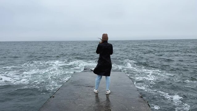 Back view of girl standing alone on pier near the sea in a storm. Woman standing near storm waves hitting the pier.