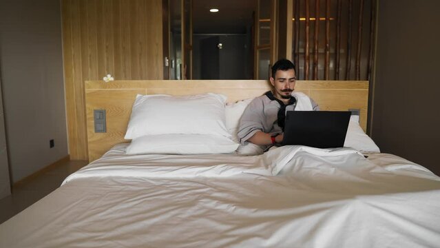 Caucasian man lying in bed is using laptop surfing in the internet then closing it and stretching. Male person works from house on computer. He is cheerful because of productive day. Freelance theme