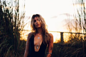 Fashion portrait of a young tanned tattooed woman in a summer dress posing in nature at sunset in...