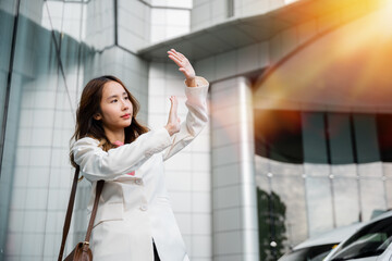 Portrait of confidence young businesswoman standing outside office building in city raise your hand to shade the sun. Happy woman wearing white suit jacket with brown bag at sunlight outdoors.