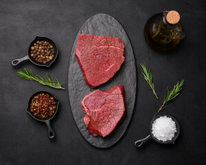 Raw piece of beef with spices pepper, rosemary sprig, salt and olive oil on a wooden board, black background.