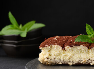 A piece of tiramisu sprinkled with cocoa, on top of a sprig of fresh mint on a black background