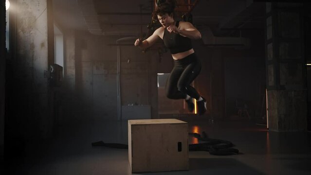 Strong female weightlifter jumps up on wooden box, develops endurance in dark smokey gym interior