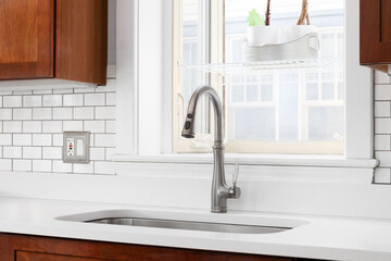 A kitchen sink detail in front of a window with a white marble countertop, white subway tile...