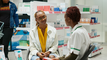 Obraz premium African american specialist showing pack of medicine to young customer, recommending pharmaceutical products in drugstore. Women talking about medicaments and vitamins in pharmacy.
