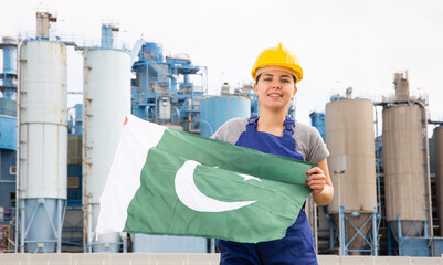 Positive young female worker in helmet waving national flag of Pakistan while standing in front of...