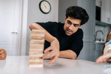 Portrait of skilled concentrated young man carefully removing wooden cube from tower structure while enjoying fun leisure time activity. Cheerful people having fun and enjoying competition.
