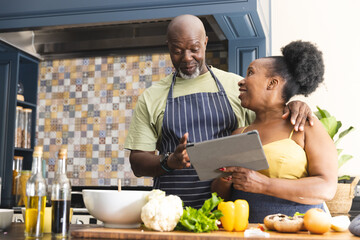 Happy senior african american couple wearing aprons and cooking with tablet in kitchen