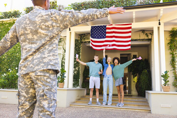 Happy diverse group of friends with usa flags, greeting biracial male american soldier outside house