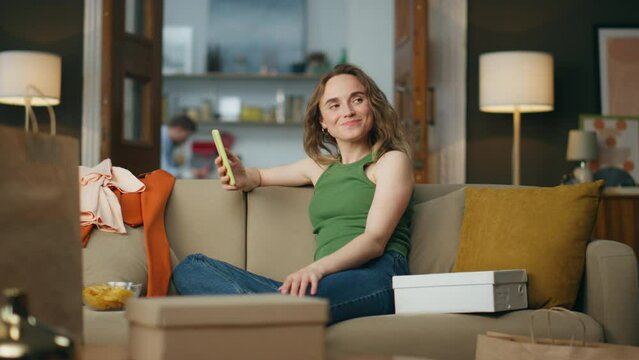 Satisfied shopaholic holding cellphone at messy house. Carefree woman thinking