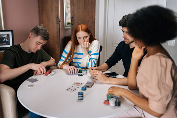 Portrait of pensive multiethnic friends having home party on weekends playing poker game in living room sitting at table. Diverse men and women satisfied with spending leisure together playing poker.