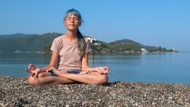 Teen enjoy sea in yoga pose. A view of young girl in yoga pose sits on the pebbles empty shore against turquoise sea waves.