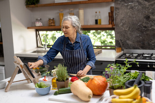 Happy senior caucasian woman cooking and using tablet in kitchen