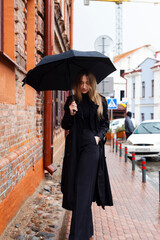 Portrait of a lovely girl in black, dressed in coat and holds an umbrella, Going down the street against a bright orange brick wall in a rainy day.