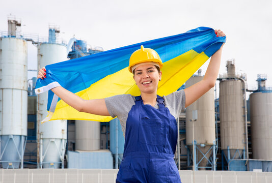 Positive young female worker in helmet waving national flag of Ukraine while standing in front of big tanks at refinery factory 