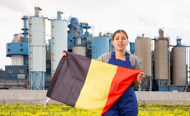 Sad young woman worker standing with big flag of Belgium against background of factory