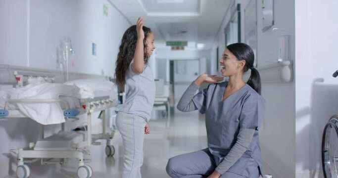 Diverse female nurse and child patient high-fiving in corridor at hospital, in slow motion