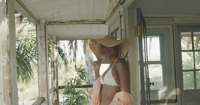 Portrait of biracial woman in bikini and sunhat smiling on porch of wooden beach house, slow motion