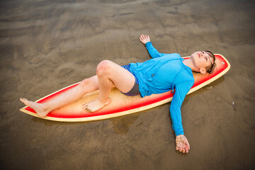 Young preteen boy on wet sand beach lying face up on surfboard with his arms outstretched