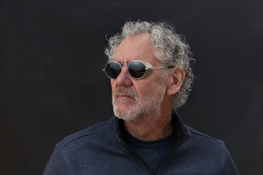Color portrait of an attractive older Caucasian man with grey hair and beard, wearing sunglasses. Looking to the side with a serious expression. Low key, dark background.