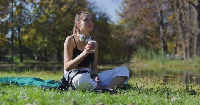 Beautiful girl drinking from a recyclable coffee cup. Fashionable young lady sitting on a field in the park.