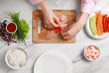 Making delicious spring rolls. Woman wrapping fresh vegetables and shrimps into rice paper at white marble table, top view