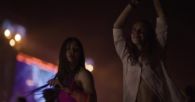 Two female friends dancing together on open space party with flashing lights. Happy beautiful girls with led rave lights on their hands. Out of focus crowd and big stage in the background.