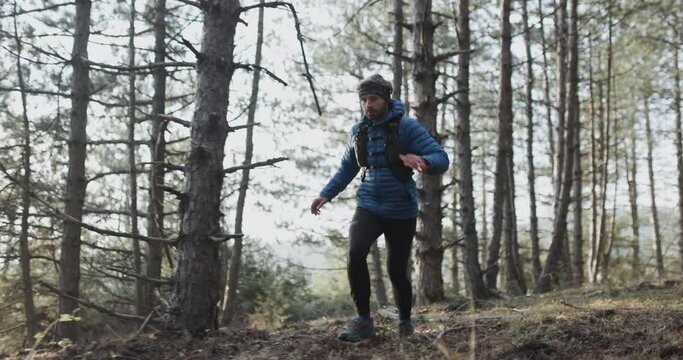 Trail runner in the woods running on a forest path and jumping. Man wearing winter sports outfit.  Sun shining between trees. Recorded at 120 fps.
