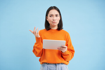 Woman with digital tablet, looks confused, cant understand smth on her gadget, stands puzzled against blue background - obrazy, fototapety, plakaty