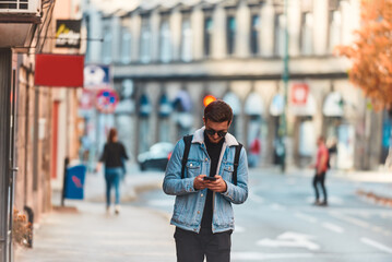 A student walking through the city carrying a backpack and wearing sunglasses while using a smartphone, representing the modern urban lifestyle and reliance on technology for communication and