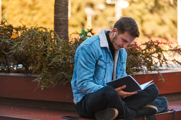 A man sitting on a bench in the city center reading a book while wearing wireless headphones,...