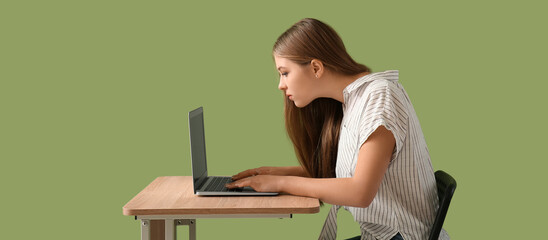 Teenage girl with bad posture using laptop while sitting at table against green background - obrazy, fototapety, plakaty