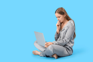 Fototapeta premium Young woman in pajamas with laptop biting nails on blue background