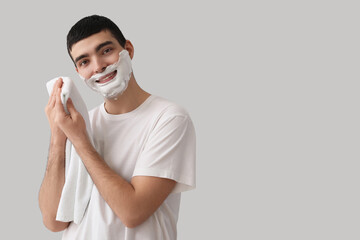 Young man wiping shaving foam from face against light background